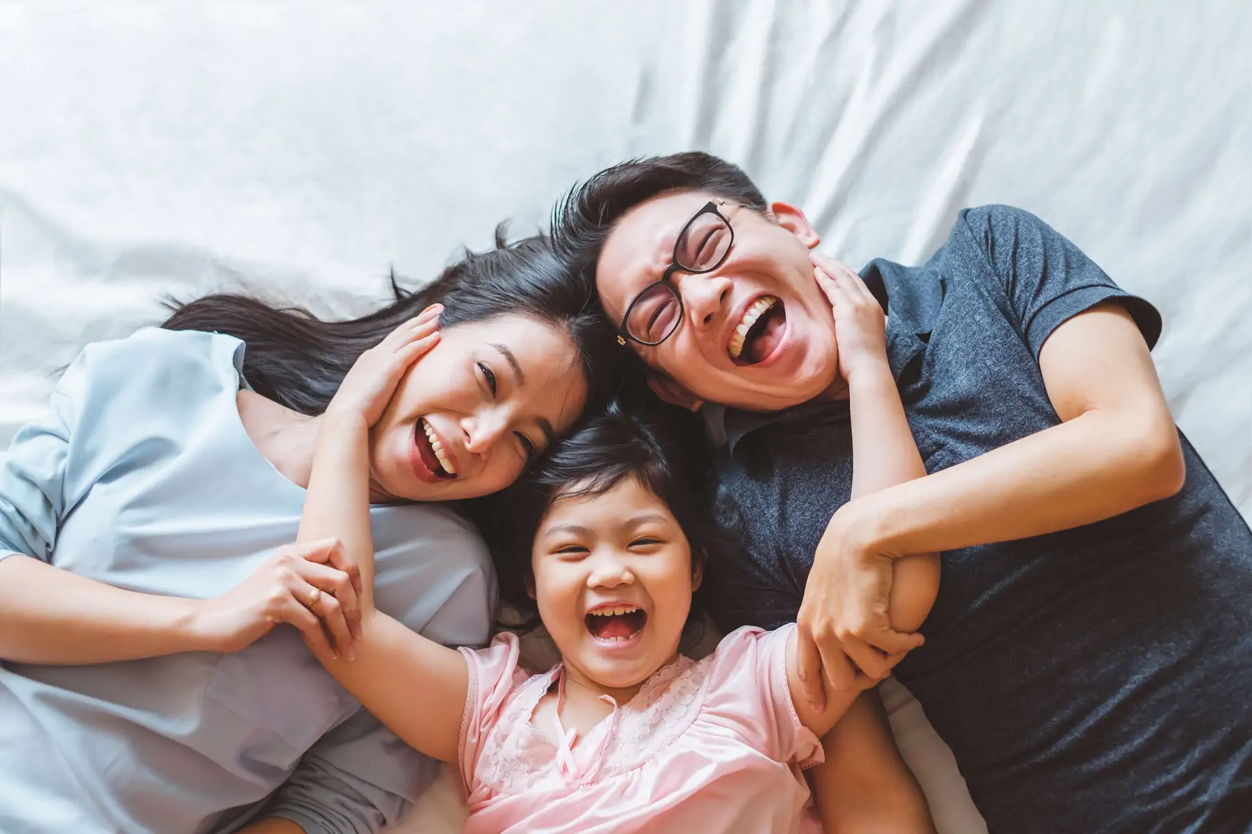 Asian mom and dad with daughter laughing and smiling while lying on a bed.